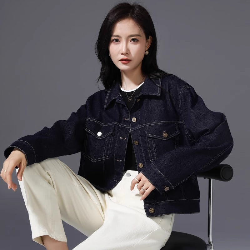 A woman wearing a dark navy blue cropped denim jacket over a black top and cream-colored wide-leg pants, seated on a black chair against a solid gray studio background.