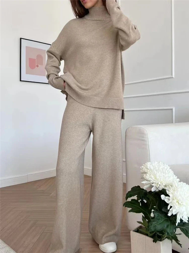 Person wearing a beige knit sweater and pants in a room with a white wall and a plant.
