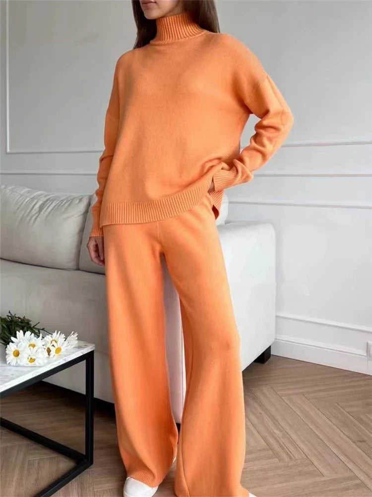A woman wearing a matching knit two-piece outfit in a warm orange color, featuring a mock-neck long-sleeve sweater and wide-leg trousers, standing in a modern living room setting.