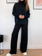 A woman wearing a black ribbed knit turtleneck sweater and matching wide-leg pants, paired with white sneakers, standing in a room with a white wall and light wood floor.