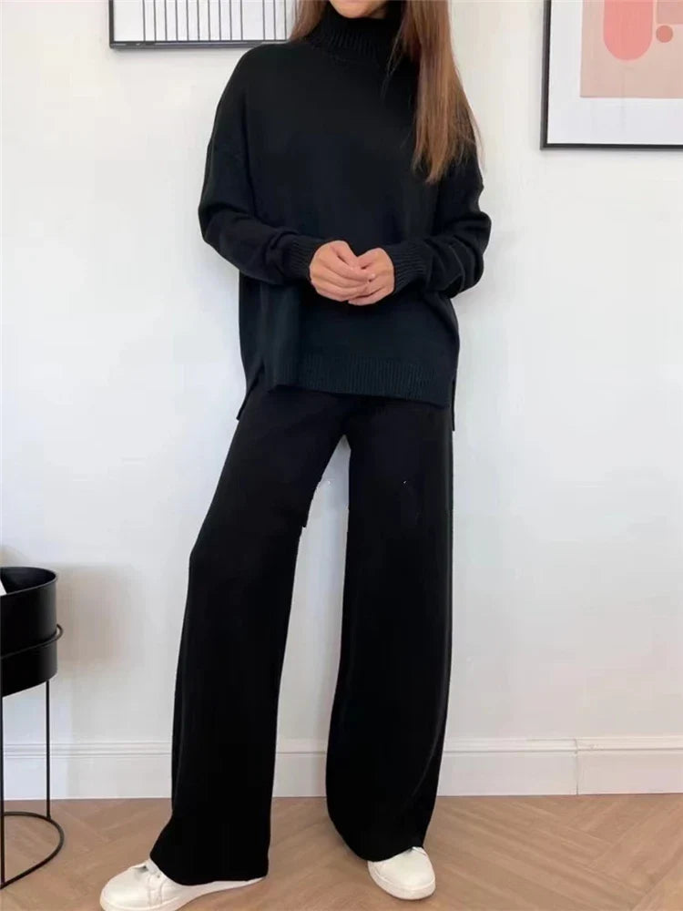 A woman wearing a black ribbed knit turtleneck sweater and matching wide-leg pants, paired with white sneakers, standing in a room with a white wall and light wood floor.