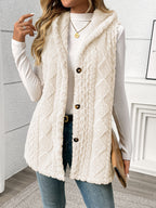A woman wearing a cream cable-knit faux-fur sleeveless vest over a white long-sleeve top and blue jeans, holding a tan clutch in a bright indoor setting.