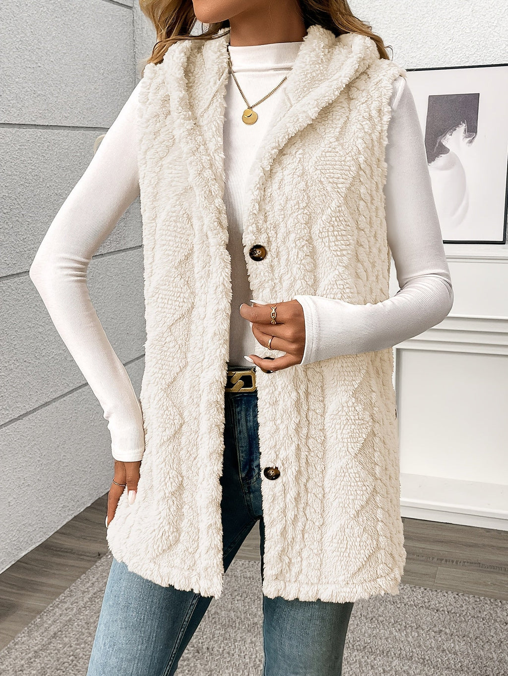 A woman wearing a cream cable-knit faux-fur vest over a white long-sleeve top and blue jeans, standing indoors against a light gray wall.
