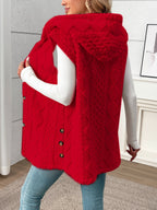 A woman wearing a bright red cable-knit hooded sleeveless vest over a white long-sleeve top and blue jeans, standing in an indoor room with light gray paneled walls.