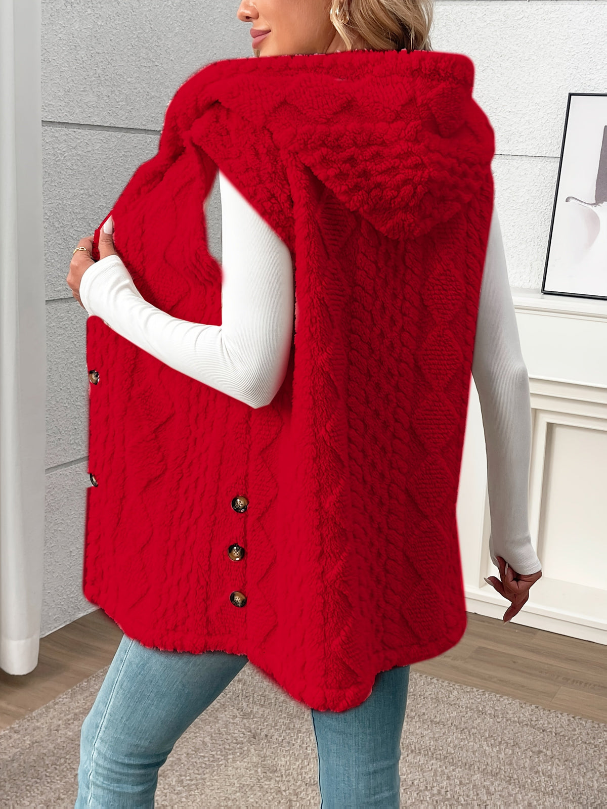A woman wearing a bright red cable-knit hooded sleeveless vest over a white long-sleeve top and blue jeans, standing in an indoor room with light gray paneled walls.
