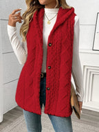 A woman wearing a bright red cable-knit hooded vest over a white long-sleeve top and blue jeans, holding a tan clutch in an indoor setting.