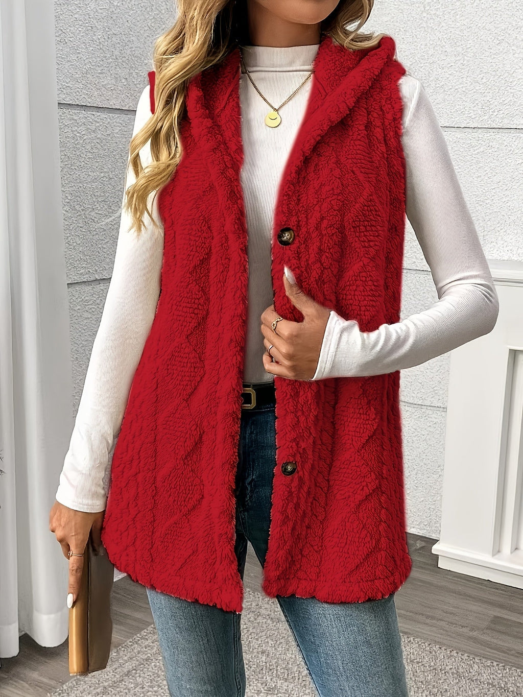 A woman wearing a bright red cable-knit sleeveless vest over a white long-sleeve top and blue jeans, standing indoors against a light gray paneled wall.