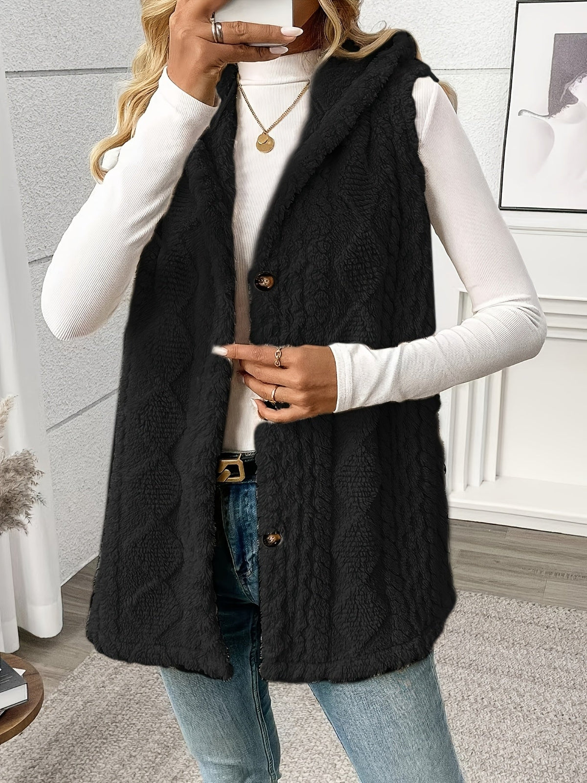 Person wearing a black knitted vest over a white long-sleeve shirt and jeans indoors.