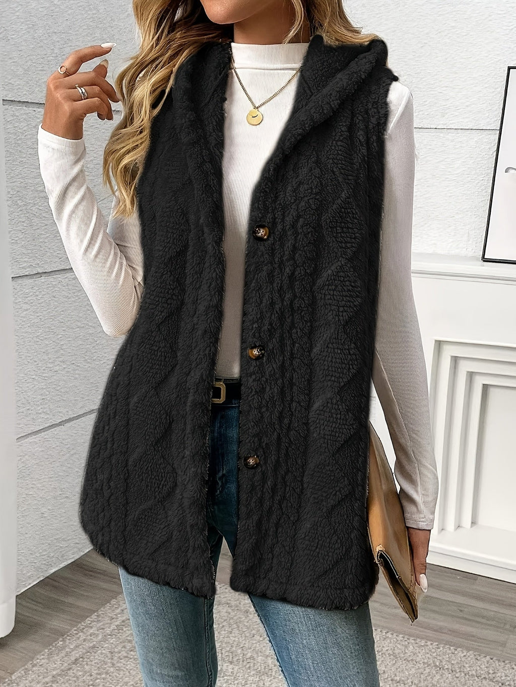 A woman wearing a black cable-knit sleeveless hooded vest over a white long-sleeve top and blue jeans, holding a tan clutch in an indoor setting.