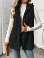 A woman wearing a black cable-knit sleeveless vest over a white long-sleeve top and blue jeans, standing indoors against a light gray paneled wall.