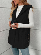 A woman wearing a black sleeveless quilted vest over a white long-sleeve top and blue jeans, holding a takeaway coffee cup against a light gray textured wall.