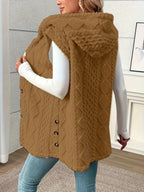 A woman wearing a brown cable-knit faux fur vest over a white long-sleeve top and blue jeans, standing in an indoor room with a textured gray wall and light wood floor.