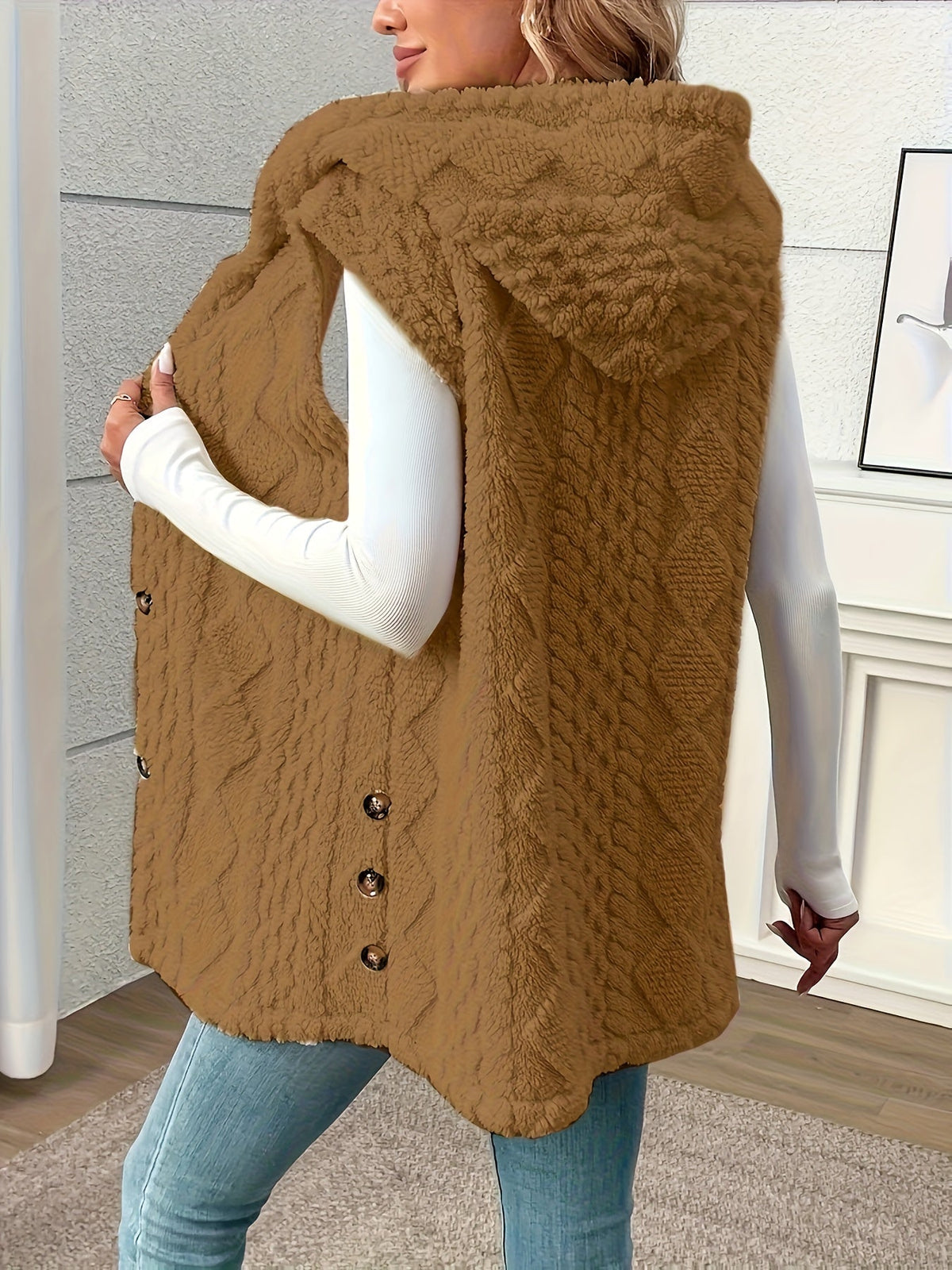 A woman wearing a brown cable-knit faux fur vest over a white long-sleeve top and blue jeans, standing in an indoor room with a textured gray wall and light wood floor.