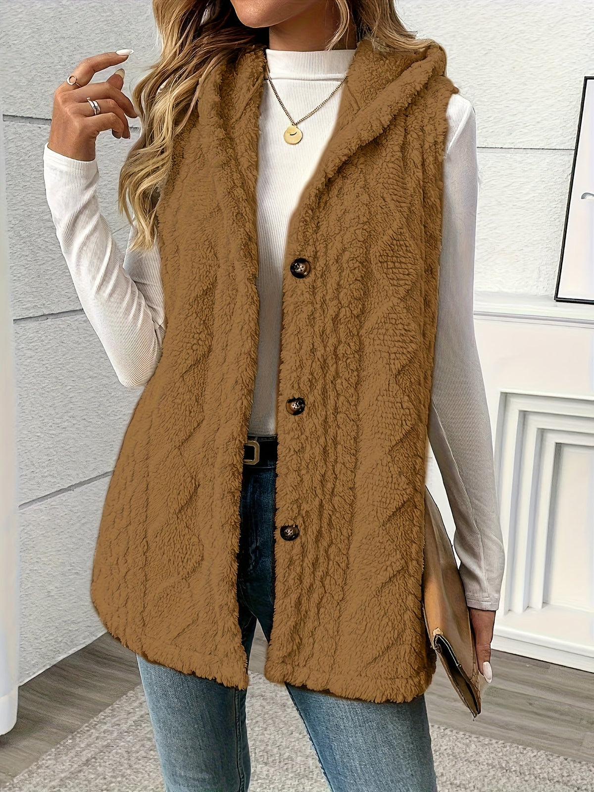 A woman wearing a brown cable-knit hooded sleeveless vest over a white long-sleeve top and blue jeans, standing indoors against a light gray wall.