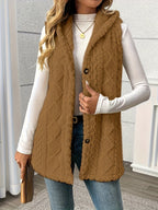 A woman wearing a brown cable-knit hooded sleeveless vest over a white long-sleeve top and blue jeans, standing indoors against a light gray wall.