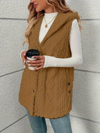 A woman wearing a brown cable-knit sleeveless vest over a white long-sleeve top and blue jeans, holding a coffee cup against a light gray textured wall.
