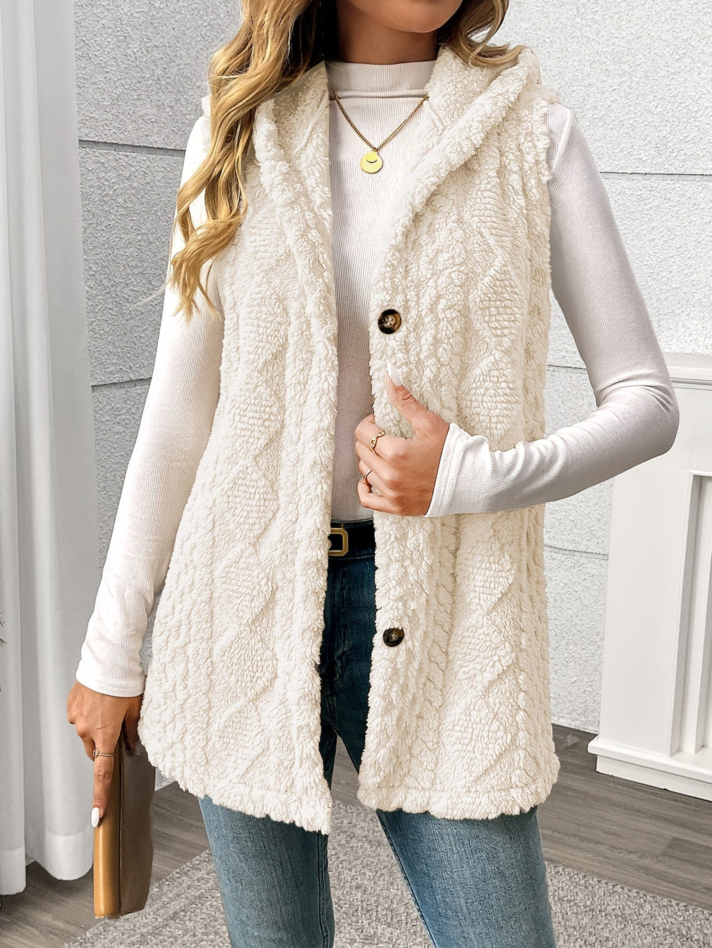 A woman wearing a cream cable-knit faux-fur vest over a white long-sleeve top and blue jeans, standing indoors against a light gray textured wall.