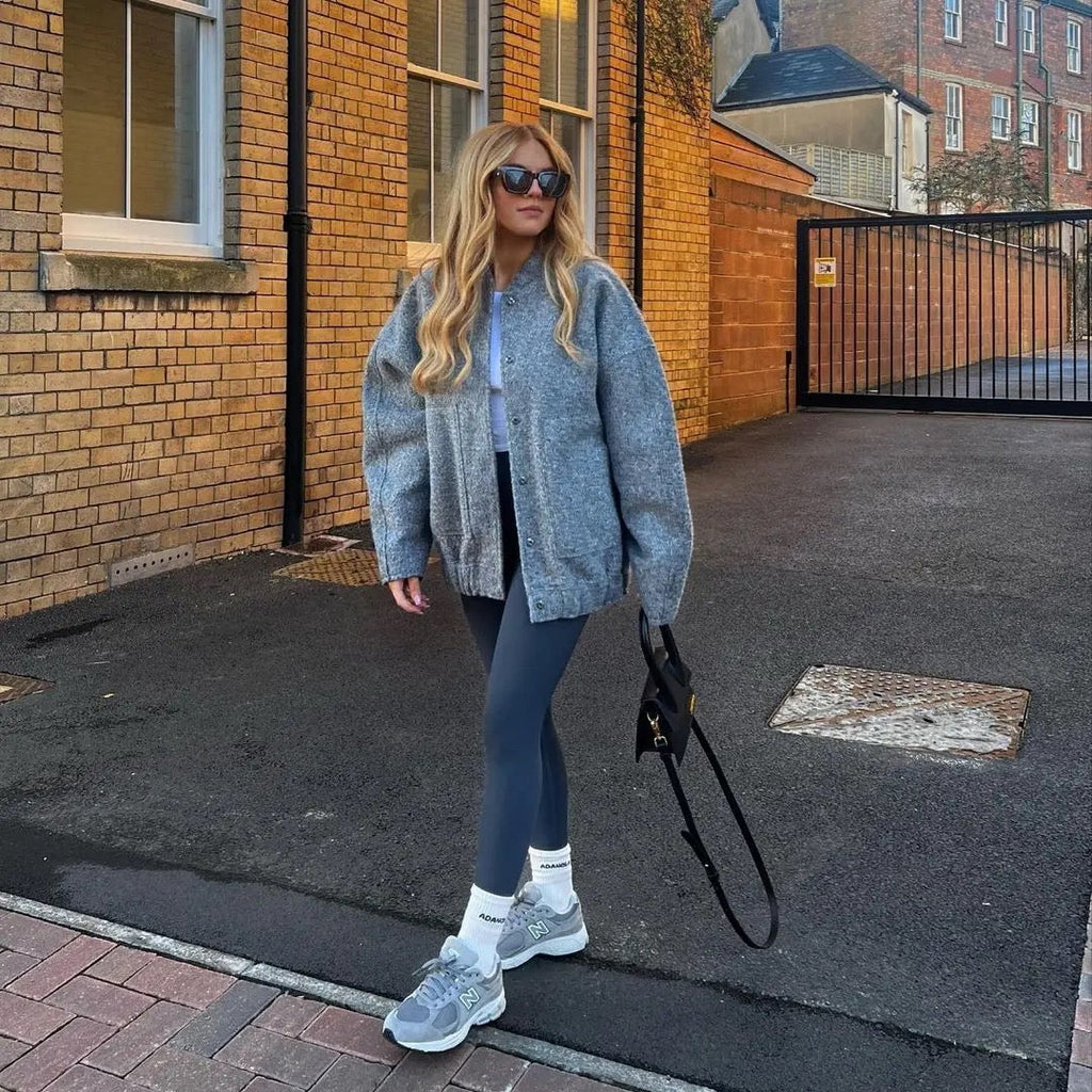 Woman in gray sweater and leggings standing on a sidewalk next to a brick building.