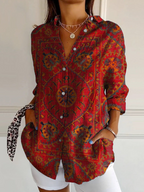 A woman wearing a red button-up shirt with a detailed multicolor bohemian paisley pattern, styled with white shorts and jewelry against a white paneled wall.