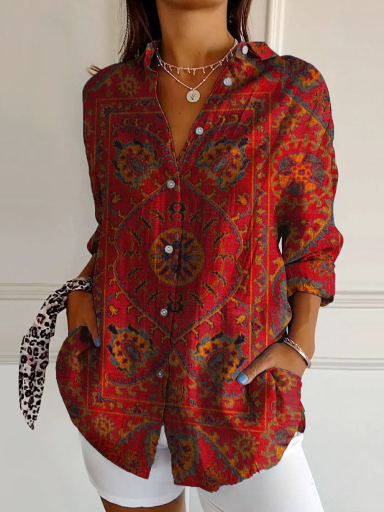 A woman wearing a red button-up shirt with a detailed multicolor bohemian paisley pattern, styled with white shorts and jewelry against a white paneled wall.