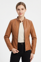 A woman modeling a tan faux-leather moto jacket with a stand collar, zippered front, and zip pockets, worn over a cream top and black pants against a light gray studio background.