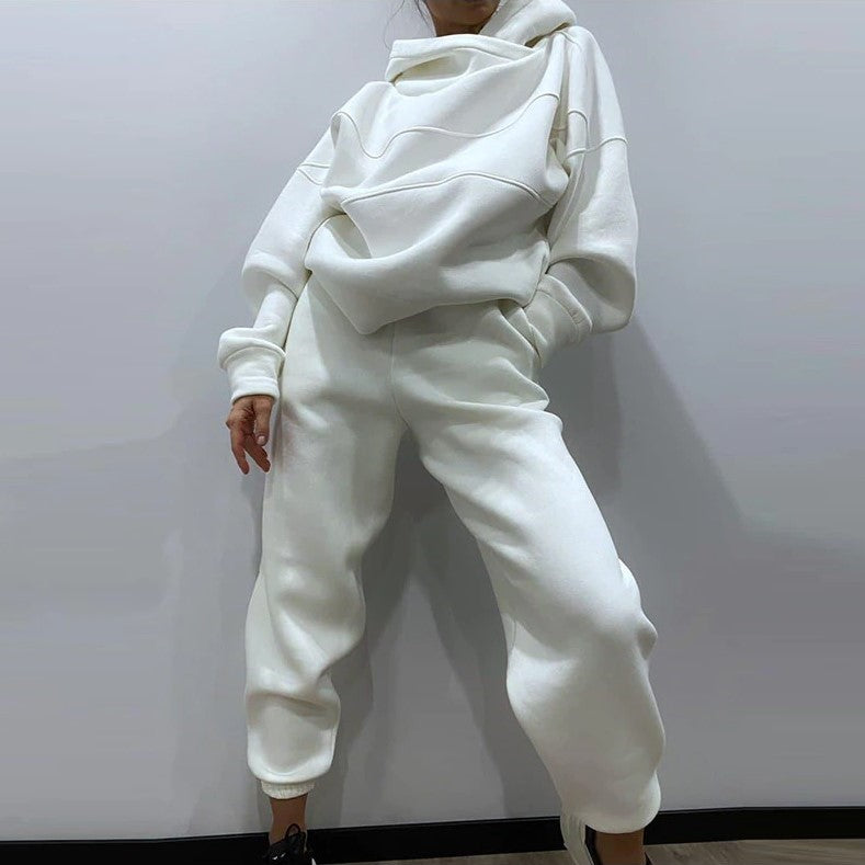 A person wearing a matching white lounge set consisting of an oversized high-neck hoodie and jogger sweatpants, standing against a plain light gray wall.