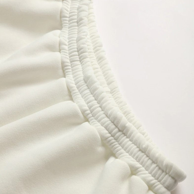 Close-up of a white or off-white pleated fabric garment detail with gathered elastic seams on a plain light background.