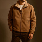 A man wearing a brown sherpa-lined zip-up fleece jacket over a white t-shirt and dark brown pants, photographed against a dark brown studio background.