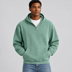 A man wearing a sage green half-zip hoodie with a kangaroo pocket, layered over a white t-shirt and blue jeans, shown from the neck down against a white background.