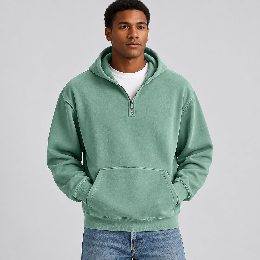 A man wearing a sage green half-zip hoodie with a kangaroo pocket, layered over a white t-shirt and blue jeans, shown from the neck down against a white background.