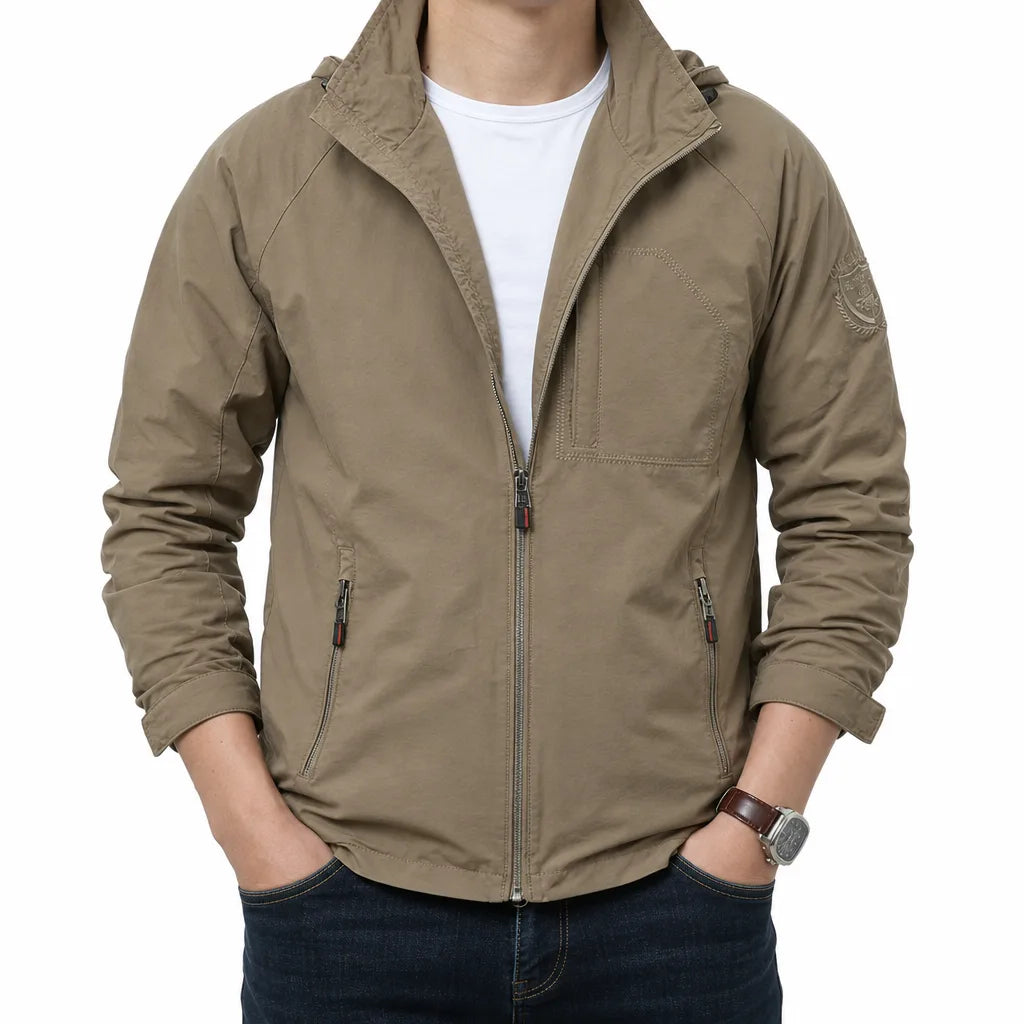 A man wearing a light brown hooded zip-up jacket over a white t-shirt and dark blue jeans, standing against a plain white background.