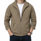 A man wearing a light brown hooded zip-up jacket over a white t-shirt and dark blue jeans, standing against a plain white background.