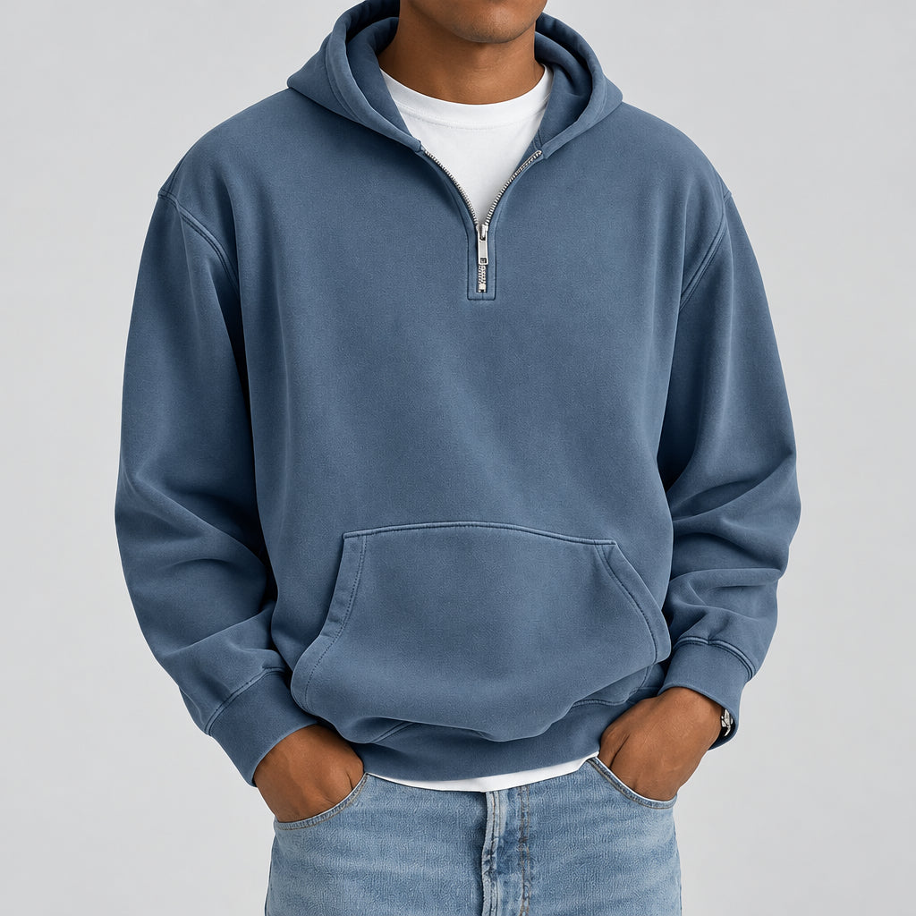 A man wearing a slate blue quarter-zip hoodie with a front kangaroo pocket, layered over a white t-shirt and blue jeans, shown from the neck down against a plain white background.